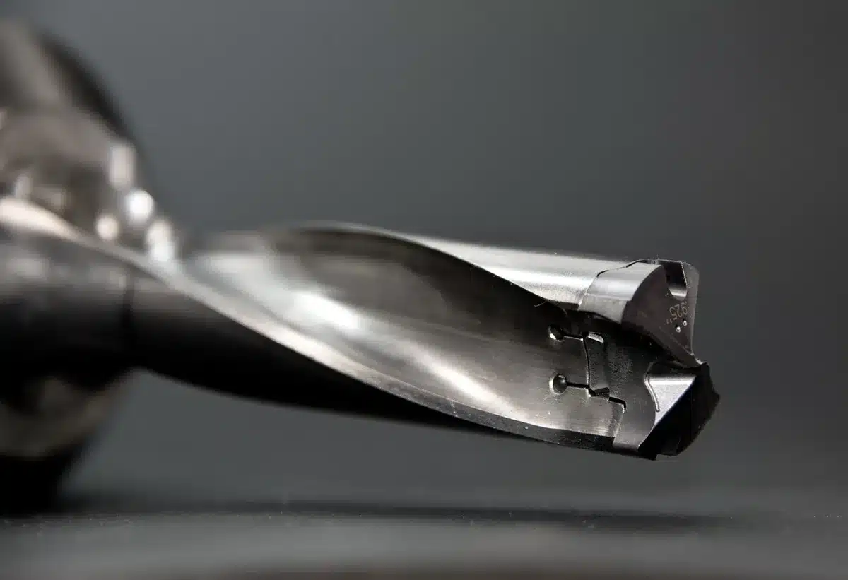An anonymous pair of hands holding a used drill over a wooden plank, drill bits and a small parts tray beside it, workshop background blurred