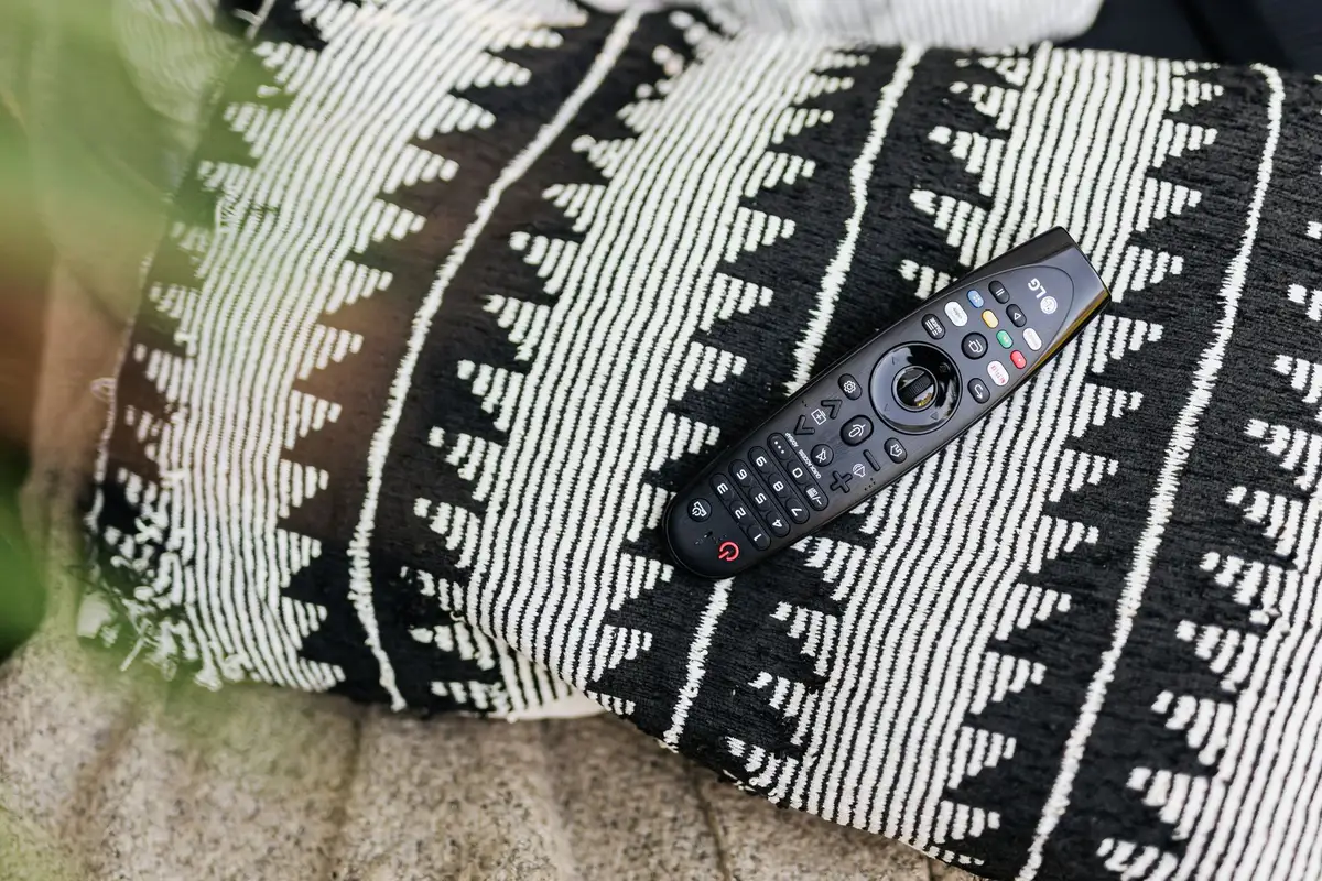 Close-up of an anonymous TV remote on a coffee table in front of a TV showing vivid colors and halo lighting on the wall, no logos or readable text; curiosity: a small price tag on the table (unreadable)