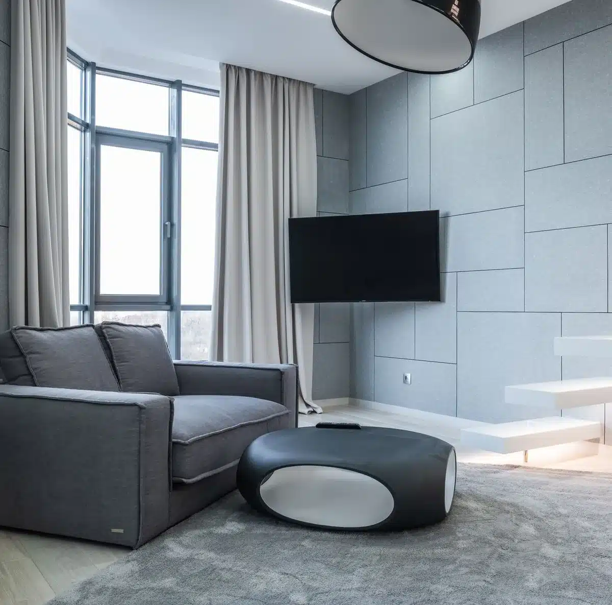Empty modern living room with a large unbranded flat-screen TV on a low console, soft natural light through a window, and a minimalist speaker beside it
